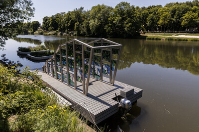 Ein Haus mit bunten Glasfenstern schwimmt auf dem Wasser. Das Kunstwerk integriert sich in die Landschaft und spiegelt sich auf der Wasseroberfläche.