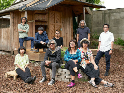 Ein Gruppenfoto von dem Kollektiv ruangrupa vor einer Holzhütte in einer Gartenumgebung.