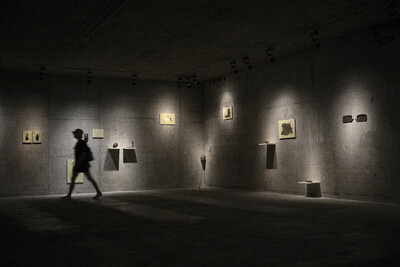 Ein Besucher bewegt sich durch einen abgedunkelten Ausstellungsraum. Wandobjekte und Bilder sind gezielt beleuchtet, wodurch eine eindrucksvolle Atmosphäre entsteht.