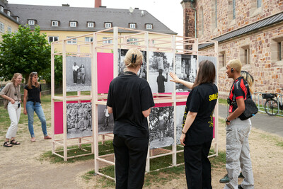 Eine Outdoor-Fotoausstellung mit mehreren Besuchenden, die gemeinsam mit einer Kunstvermittlerin die Bilder betrachten.