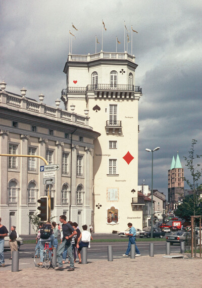 Turmartiger Anbau eines klassizistischen Gebäudes mit Spielkarten-Symbolen und Textfragmenten an der Fassade. Menschen bewegen sich auf dem Gehweg im Vordergrund. Im Hintergrund ist ein Kirchturm mit grünem Dach sichtbar.