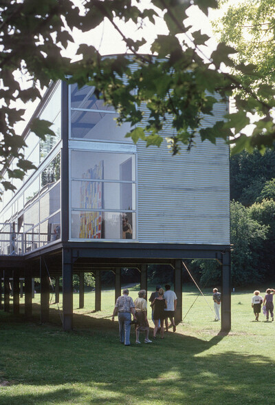 Modular wirkendes Gebäude auf Stelzen, mit großen Glasflächen und metallverkleideten Wänden. Durch das Fenster ist eine farbige Wandarbeit im Inneren zu erkennen. Mehrere Personen gehen auf das Gebäude zu.