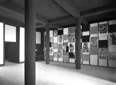 A black and white photograph of an art exhibition with modern architectural elements. The exhibition features a wall installation with a collage of images and artworks framed by black and white panels. Solid wooden columns and a raw concrete ceiling give the space an industrial feel.