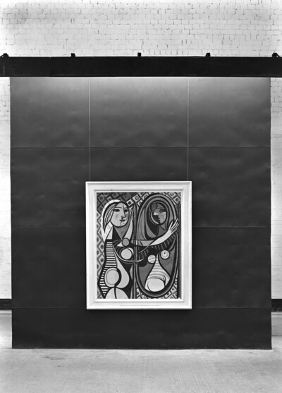 Eine Schwarz-Weiß-Fotografie eines Ausstellungsraums mit einem einzelnen Kunstwerk, das zentral an einer schwarzen Wand präsentiert wird. Das Gemälde zeigt "Mädchen vor Spiegel" von Pablo Picasso, das mit geometrischen Formen und abstrakten Mustern gestaltet ist. Die Wand im Hintergrund besteht aus weißem Backstein, während ein dunkler Balken das Bild oben rahmt.