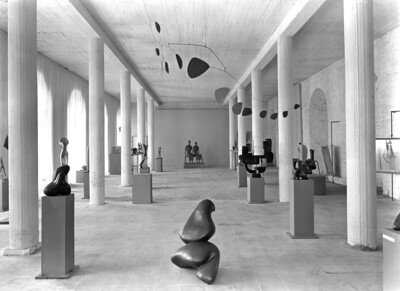 Schwarz-Weiß-Fotografie eines Ausstellungsraums der ersten documenta. Der Raum ist groß und hell, mit hohen Decken und einer schlichten, modernen Gestaltung. An den Wänden hängen mehrere Gemälde. Im Vordergrund ist eine abstrakte Skulptur auf dem Boden zu sehen, links und rechts angeordnet stehen weitere Skulpturen auf Sockeln.