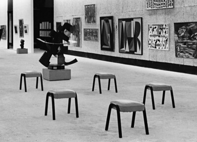 Eine schwarz-weiße Aufnahme einer Kunstausstellung mit abstrakten Gemälden an der Wand, einer modernen Metallskulptur auf einem Sockel und mehreren kleinen Hockern im Vordergrund. Die Ausstellungshalle hat einen schlichten, funktionalen Stil mit einfachen Wänden und einem offenen Raumkonzept.