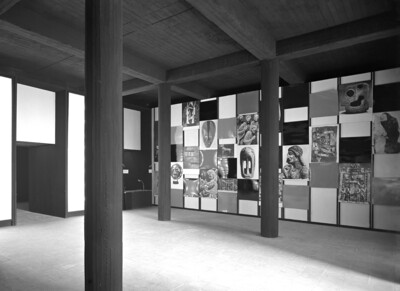 Eine Schwarz-Weiß-Fotografie einer Kunstausstellung mit modernen architektonischen Elementen. Die Ausstellung zeigt eine Wandinstallation mit einer Collage aus Bildern und Kunstwerken, die von schwarzen und weißen Paneelen umrahmt sind. Massive Holzsäulen und eine rohe Betondecke verleihen dem Raum eine industrielle Atmosphäre.