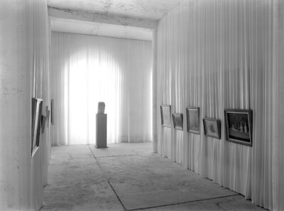 Schwarz-Weiß-Fotografie eines Ausstellungsraums der ersten documenta. Im Hintergrund steht eine Skulptur auf einem Sockel am Ende des Raumes. Links und rechts an den Wänden hängen verschiedene Gemälde, die an mit Stoff bespannten Wänden präsentiert werden.