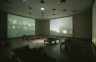 Dunkler Ausstellungsraum mit mehreren großen Videoprojektionen auf drei Wänden, die neblige nächtliche Stadtansichten zeigen. Im Raum verteilt stehen gepolsterte Holzliegen, auf denen mehrere Personen liegen und die Projektionen betrachten.