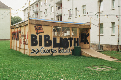 Einfacher Holzbau mit Graffiti-Schriftzug „Bibliothek Georges Bataille“ auf einer Wiese.