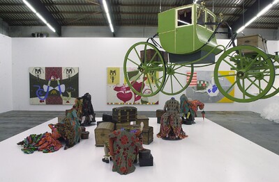 Installation mit kopflosen Figuren in bunten Stoffen, alten Koffern und einer grünen Kutsche, die von der Decke hängt.