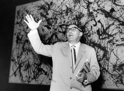 Arnold Bode steht vor einem Werk von Jackson Pollock. Eine Hand hält er angewinkelt nach oben, in der anderen Hand hält er Unterlagen.
