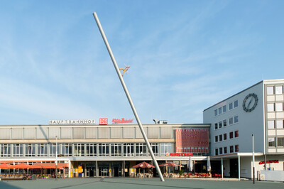 Schräge Metallskulptur mit einer Figur, die scheinbar nach oben aufsteigt, vor dem Hauptbahnhof Kassel. Im Hintergrund das Bahnhofsgebäude mit großen Fenstern, Restaurants mit roten Sonnenschirmen und eine weiße Fassade mit einer großen Uhr.