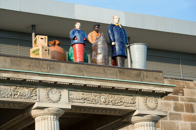 Bunte Skulpturen von Menschenfiguren und Gegenständen wie Kisten und Säcken stehen auf einem klassischen Gebäudegesims mit verzierten Reliefs. Im Hintergrund ein modernes Gebäude mit grauen Fassadenelementen.