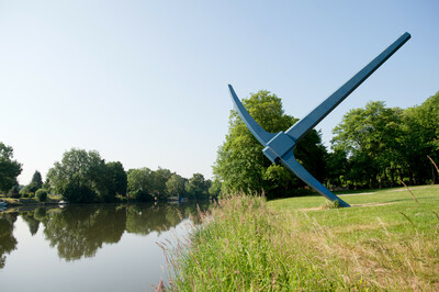 Große, blaue Spitzhacke als Skulptur, die mit dem Stiel schräg in einer Wiese steckt. Die Szene befindet sich an einem Flussufer mit grünem Bewuchs und Bäumen im Hintergrund unter klarem Himmel.