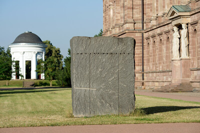 Ein grober, rechteckiger Granitblock mit sichtbaren Einschnitten und Bohrlöchern steht auf einer Rasenfläche. Im Hintergrund ein historisches Sandsteingebäude mit Karyatidenfiguren sowie ein weißer Rundtempel mit Säulen und Kuppeldach.