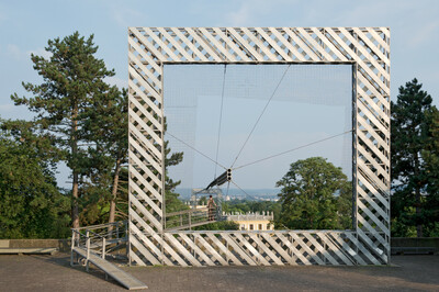 Large, square metal construction with diagonal struts that looks like a frame. In the middle is a net with a round opening from which a tubular element protrudes. In the background are trees, a classical building and a wide landscape. A person is standing on a ramp within the construction.