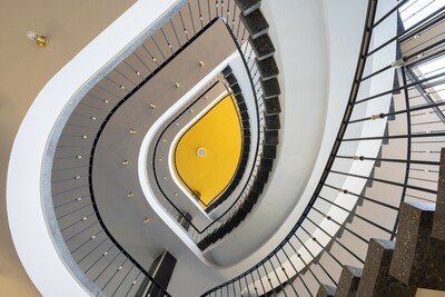Blick von unten in ein mehrstöckiges, geschwungenes Treppenhaus mit dunklen Stufen und weißen Wänden. Die Geländer mit filigranen Metallstreben verlaufen parallel zu den Treppen. Im Zentrum ein gelb gefärbter Wandbereich mit einer Deckenlampe.