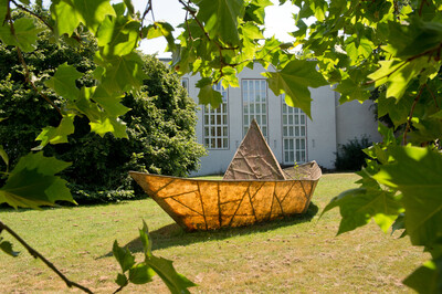 Sculpture of a large, paper boat-like ship made of metal with a transparent structure on a grassy area, framed by green leaves in the foreground. In the background, a modern building with tall windows.
