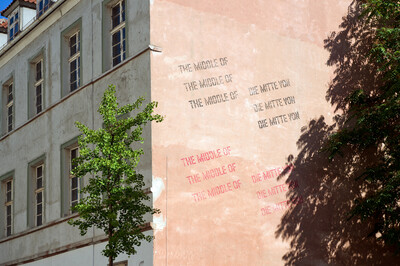 Hauswand mit verblasster rosafarbener Fassade, auf der mehrfach in Schwarz und Rot der Text „THE MIDDLE OF“ und „DIE MITTE VON“ in verschiedenen Größen aufgedruckt ist. Ein grüner Baum steht im Vordergrund, während ein weiterer Baum rechts einen Schatten auf die Wand wirft.
