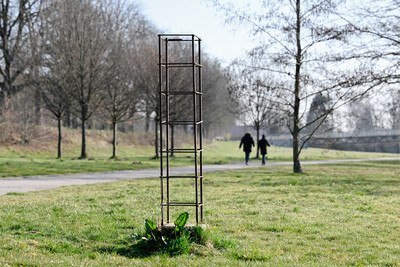 Eine schlanke, rechteckige Metallstruktur aus rostigen Bewehrungsstäben steht auf einer Wiese. Darunter wächst eine kleine Pflanze. Im Hintergrund verlaufen ein Fußweg, kahle Bäume und eine Brücke. Zwei Personen gehen in der Ferne in Richtung Horizont.