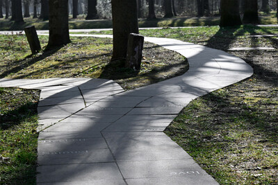 Ein geschwungener, gepflasterter Weg verläuft durch einen bewaldeten Park. Auf den Platten sind verschiedene eingravierte Wörter zu erkennen. Schatten der Bäume fallen auf den Weg und das umgebende Gras.