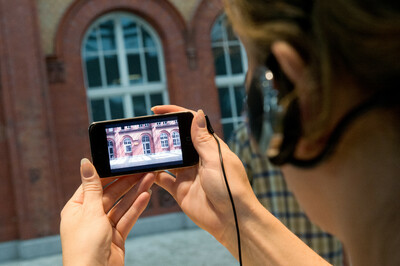 Eine Person hält ein Smartphone in beiden Händen, auf dessen Bildschirm eine rote Backsteinfassade mit Fenstern zu sehen ist. Ein Kopfhörerkabel ist mit dem Gerät verbunden. Im Hintergrund ist unscharf eine ähnliche Fassade eines Gebäudes zu erkennen.