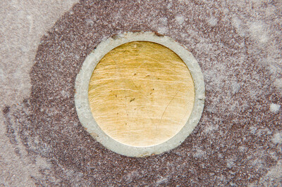 Close-up of a circular, golden metal disc embedded flush in a stone slab. The surface shows fine scratches and signs of use.