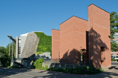 Backstein-Skulptur mit mehreren rechteckigen Türmen und offenen Durchgängen, umgeben von einem schwarzen Geländer. Im Hintergrund ein modernes Gebäude mit einer geneigten, teilweise begrünten Fassade. Der Himmel ist klar und blau, mit Schatten von Bäumen auf der Skulptur.
