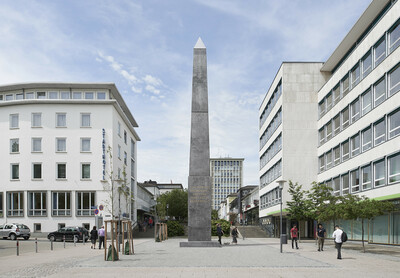 Großer, dunkler Obelisk mit einer goldenen Inschrift auf einer städtischen Platzfläche, umgeben von modernen Gebäuden mit vielen Fenstern. Die Inschrift lautet: „ICH WAR EIN FREMDLING UND IHR HABT MICH BEHERBERGT.“ Mehrere Menschen gehen oder stehen um das Kunstwerk herum, einige Bäume säumen den Platz.