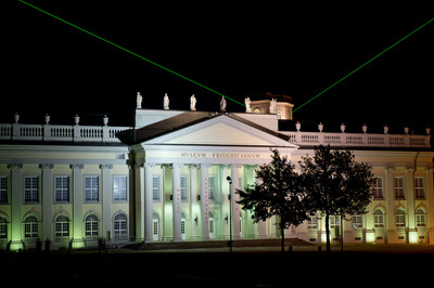 Beleuchtetes Museum Fridericianum bei Nacht mit grünen Laserstrahlen, die von der Dachmitte schräg in den Himmel ragen. Im Vordergrund stehen Bäume, die teilweise die Fassade verdecken.